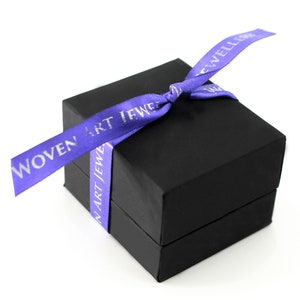 May include: A black gift box with a purple ribbon tied around it. The ribbon has the text "WOVEN ART JEWELRY" printed on it in silver lettering.