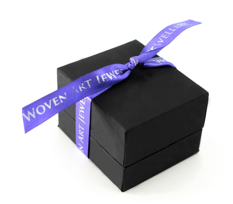 May include: A black gift box with a purple ribbon tied around it. The ribbon has the text "WOVEN ART JEWELRY" printed on it in silver lettering.