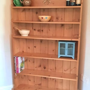 May include: A light brown wooden dresser with several shelves. The top shelf displays wine bottles. Other shelves hold a green ceramic jug, a small blue cabinet, a wooden bowl, and cookbooks. The dresser is constructed from light-coloured wood.