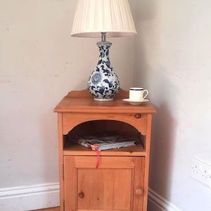 May include: A wooden bedside table with a lamp and a teacup. The lamp features a cream shade and a blue and white patterned base. A book with a red bookmark rests on the shelf below.