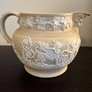 Puede incluir: Una jarra de cerámica beige con asa y boquilla. La jarra está decorada con diseños en relieve blancos de uvas, hojas y criaturas míticas. La jarra tiene una base redonda y una abertura ancha.