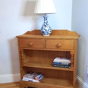 May include: A light brown wooden bookcase with two drawers and two shelves. A white lampshade sits atop a blue and white patterned lamp. Books and magazines are on the shelves.