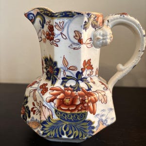 Könnte beinhalten: Eine weiße Keramikkrug mit einem Blumenmuster in Rot, Blau und Orange. Der Krug hat eine einzigartige, eckige Form und einen gebogenen Griff. Das Blumenmuster umfasst große Blüten und detailliertes Laub, was einen Vintage-Look erzeugt.