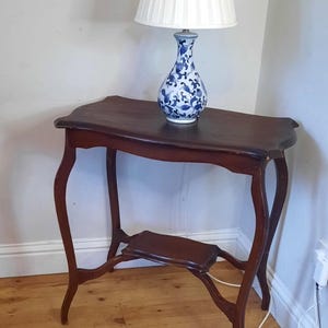 May include: An antique dark brown wooden side table with curved legs and a lower shelf. A white lampshade sits on a blue and white patterned vase. The table is on a wooden floor. The table's height is approximately 76 cm.