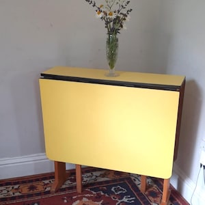 May include: A yellow drop-leaf table with wooden legs, holding a clear glass vase filled with white and purple flowers. The table is set on a patterned red rug. The table is approximately 76 cm long.