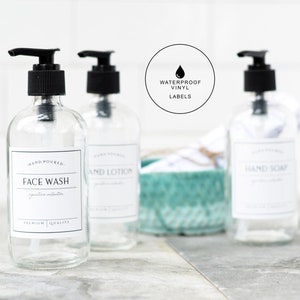 May include: Three clear glass bottles with black pump tops and white labels. The labels read "Hand Poured Face Wash", "Hand Poured Hand Lotion", and "Hand Poured Hand Soap". The bottles are sitting on a light gray surface. A teal basket with white towels is in the background. A black circle with a white water drop and the text "Waterproof Vinyl Labels" is in the upper right corner of the image.