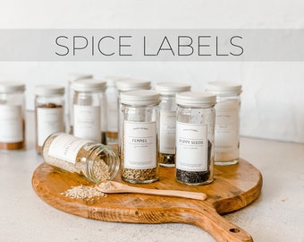 May include: A wooden cutting board with a variety of glass spice jars with white labels. The labels have black text and include the names "Fennel", "Poppy Seeds", and "Salt".