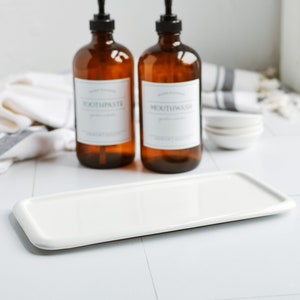 May include: Two amber glass bottles with pumps, one labelled "Toothpaste" and the other "Mouthwash", sitting on a white rectangular ceramic tray. The bottles are on a white tiled surface with a white and grey striped towel in the background.