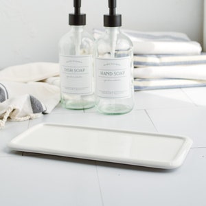 May include: Two glass bottles with black pump tops and labels that read "Hand Poured Dish Soap" and "Hand Poured Hand Soap". The bottles are sitting on a white rectangular tray.