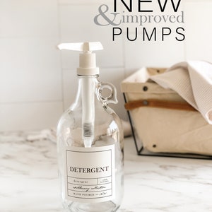 May include: A clear glass bottle with a white pump dispenser and a black and white label that reads "DETERGENT, detergent, apothecary collection, HAND POURED - NYC". The bottle is sitting on a white marble surface.