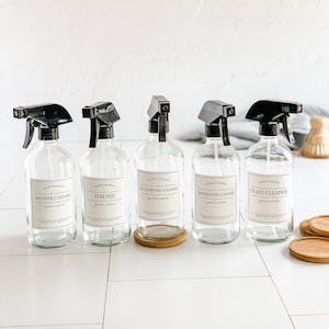 May include: Five clear glass spray bottles with black trigger tops and white labels. The labels have the words "Hand Poured" and the names of different cleaning solutions: "Shower Cleaner", "Thieves", "All Purpose Cleaner", "Bathroom Cleaner", and "Glass Cleaner".