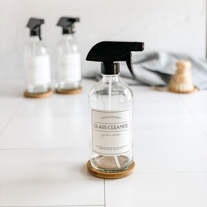 May include: Three glass spray bottles with black trigger sprayers. The center bottle has a white label that reads "Hand Poured. Glass Cleaner. Signature Collection. Premium Quality."