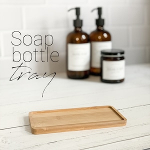 May include: A rectangular wooden tray with a natural wood finish. The tray is empty and is sitting on a white surface. The tray is in focus, while the background is blurred. The background includes two glass bottles with pumps and a small glass jar. The bottles and jar are out of focus. The text "Soap bottle tray" is visible in the image.