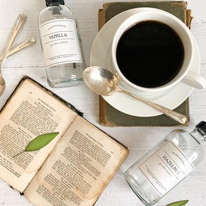 May include: Two glass bottles with labels reading "HAND POURED" and "VANILLA" and "HAZELNUT" with the text "signature scent" and "premium quality" on a white surface with a cup of coffee, a book, and green leaves.