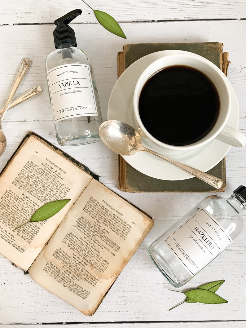 May include: Two glass bottles with labels reading "HAND POURED" and "VANILLA" and "HAZELNUT" with the text "signature scent" and "premium quality" on a white surface with a cup of coffee, a book, and green leaves.