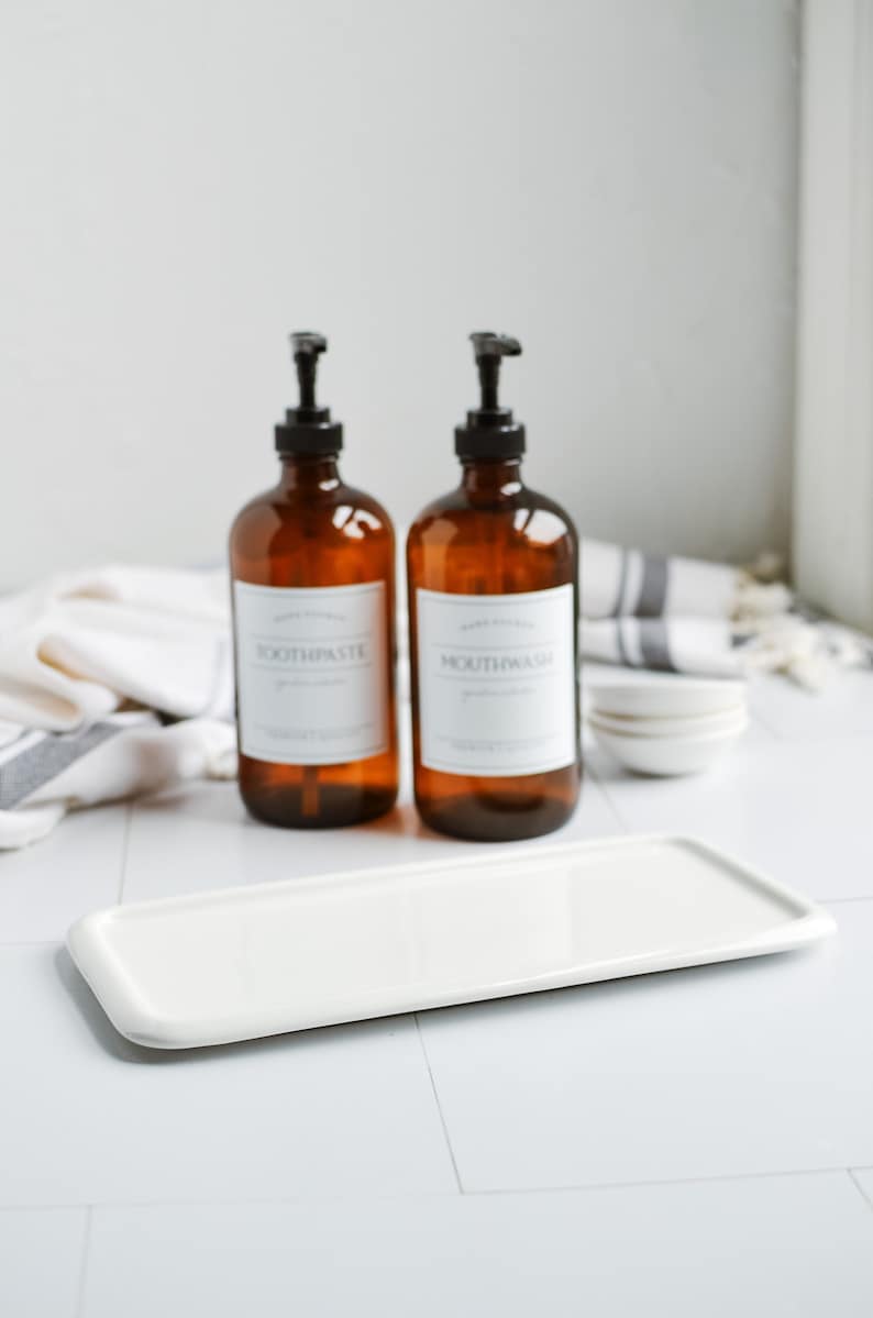 May include: Two amber glass bottles with pumps, one labelled "Toothpaste" and the other "Mouthwash", sitting on a white rectangular ceramic tray. The bottles are on a white tiled surface with a white and grey striped towel in the background.