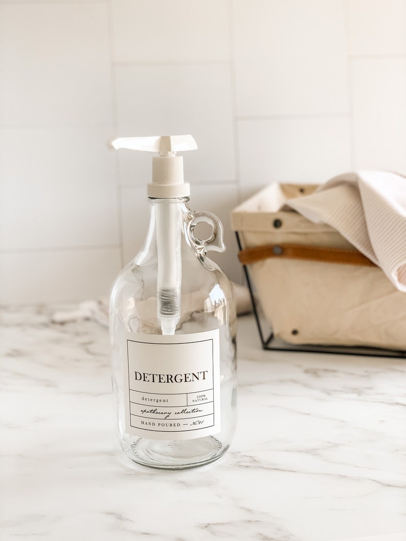 May include: A clear glass bottle with a white pump dispenser and a label that reads "DETERGENT" with the text "detergent", "100% NATURAL", "apothecary collection", and "HAND POURED - NYC".