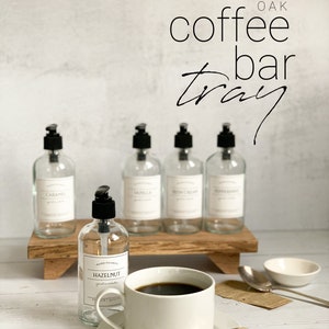 May include: White oak coffee bar tray with five glass bottles with black pump tops and labels. The bottles are filled with liquids. The tray is sitting on a white wooden surface. A white coffee cup with a saucer and a silver spoon is in the foreground. A small white bowl is on the right side of the image.