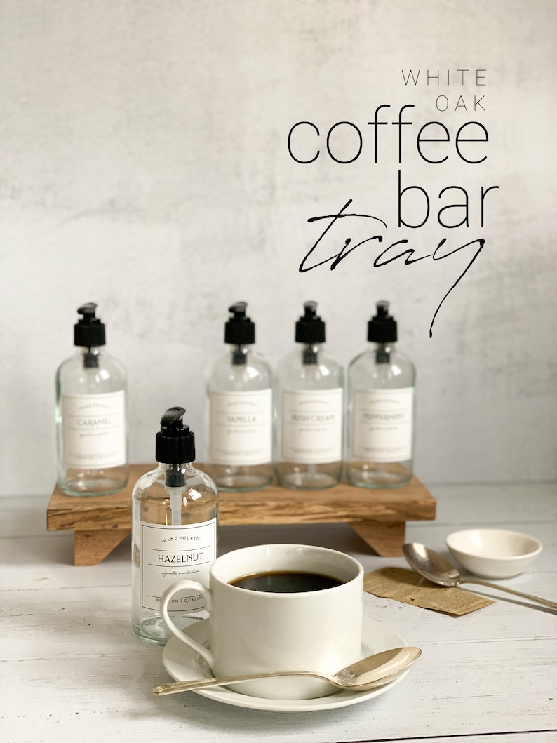 May include: White oak coffee bar tray with five glass bottles with black pump tops and labels. The bottles are filled with liquids. The tray is sitting on a white wooden surface. A white coffee cup with a saucer and a silver spoon is in the foreground. A small white bowl is on the right side of the image.