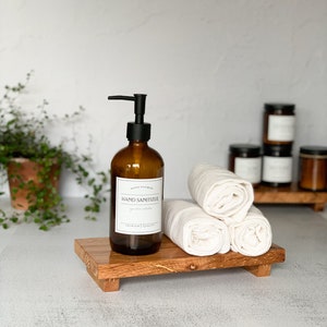 May include: A brown glass hand sanitizer bottle with a black pump dispenser and a white label that reads "Hand Sanitizer". The bottle sits on a small, rectangular wooden tray with three rolled white towels. Several jars are in the background.