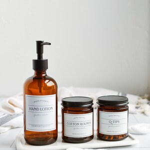 May include: Three amber glass bottles with black lids and labels. The bottles are on a white tray. The labels read "Hand Lotion", "Cotton Rounds", and "Q-Tips".