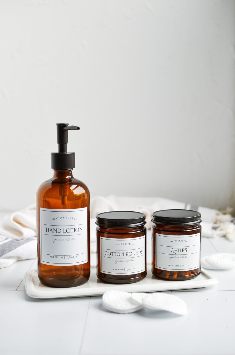May include: Three amber glass bottles with black lids and labels. The bottles are on a white tray. The labels read "Hand Lotion", "Cotton Rounds", and "Q-Tips".