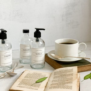 May include: Three clear glass bottles with black pump tops and labels. The labels read "Hand Poured Caramel", "Hand Poured Hazelnut", and "Hand Poured Vanilla". The bottles are on a white surface with a book, a cup of coffee, and spoons.