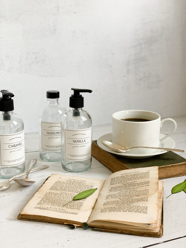 May include: Three clear glass bottles with black pump tops and labels. The labels read "Hand Poured Caramel", "Hand Poured Hazelnut", and "Hand Poured Vanilla". The bottles are on a white surface with a book, a cup of coffee, and spoons.