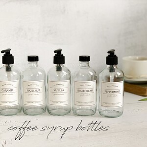 May include: Five clear glass bottles with black pump tops and labels. The labels are white with black text and say "Hand Poured" and the flavor of the syrup inside. The flavors are Caramel, Hazelnut, Vanilla, Irish Cream, and Peppermint. The bottles are sitting on a white surface with the text "coffee syrup bottles" written below them.