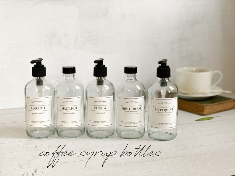 May include: Five clear glass bottles with black pump tops and labels. The labels are white with black text and say "Hand Poured" and the flavor of the syrup inside. The flavors are Caramel, Hazelnut, Vanilla, Irish Cream, and Peppermint. The bottles are sitting on a white surface with the text "coffee syrup bottles" written below them.