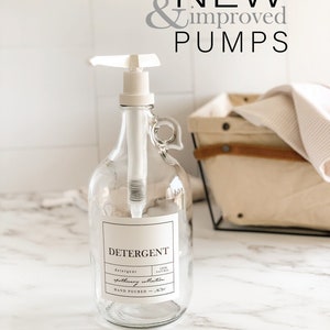 May include: A clear glass bottle with a white pump dispenser and a label that reads "DETERGENT" with the text "100% NATURAL" and "gathering collection" underneath. The bottle is sitting on a white marble countertop.