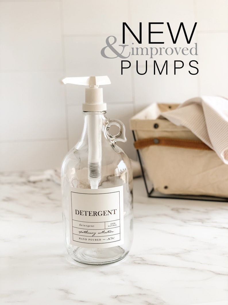 May include: A clear glass bottle with a white pump dispenser and a label that reads "DETERGENT" with the text "100% NATURAL" and "gathering collection" underneath. The bottle is sitting on a white marble countertop.