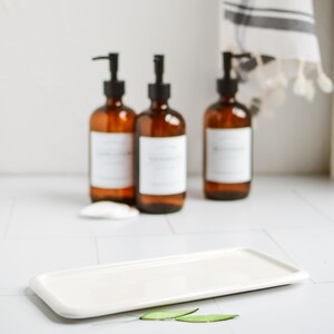 May include: Three amber glass bottles with black pump tops and white labels sit on a white countertop. A white rectangular ceramic dish sits in front of the bottles with two green leaves on the countertop.