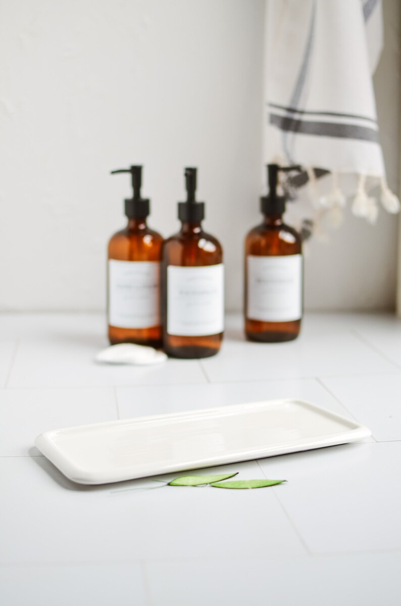 May include: Three amber glass bottles with black pump tops and white labels sit on a white countertop. A white rectangular ceramic dish sits in front of the bottles with two green leaves on the countertop.