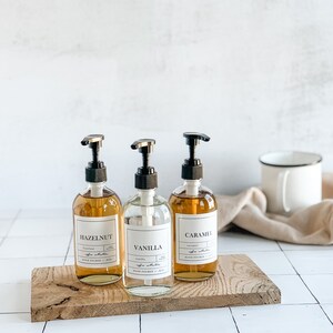 Puede incluir: Tres botellas de vidrio con bombas, etiquetadas "Hazelnut", "Vanilla" y "Caramel", sentadas sobre una tabla de madera. Las botellas est&aacute;n llenas de un l&iacute;quido marr&oacute;n claro.