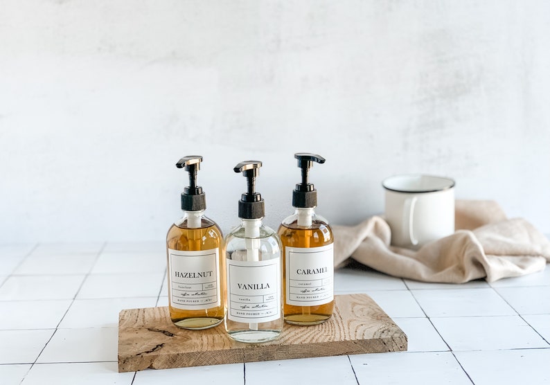 Puede incluir: Tres botellas de vidrio con bombas, etiquetadas "Hazelnut", "Vanilla" y "Caramel", sentadas sobre una tabla de madera. Las botellas est&aacute;n llenas de un l&iacute;quido marr&oacute;n claro.