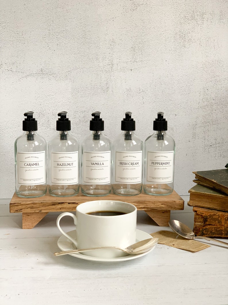 May include: Five clear glass bottles with black pump tops and labels. The labels are white with black text and list the following scents: Caramel, Hazelnut, Vanilla, Irish Cream, and Peppermint. The bottles are arranged on a wooden shelf with a white cup of coffee and a saucer on the shelf below.