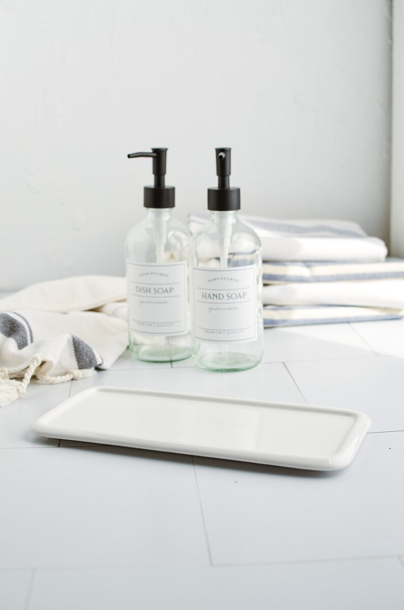 May include: Two glass bottles with black pump tops and labels that read "Hand Poured Dish Soap" and "Hand Poured Hand Soap". The bottles are sitting on a white rectangular tray.