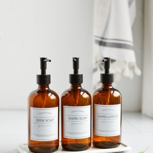 May include: Three amber glass bottles with black pump tops and white labels. The labels read "Hand Poured", "Dish Soap", "Hand Soap", and "Hand Lotion". The bottles are sitting on a white tray.