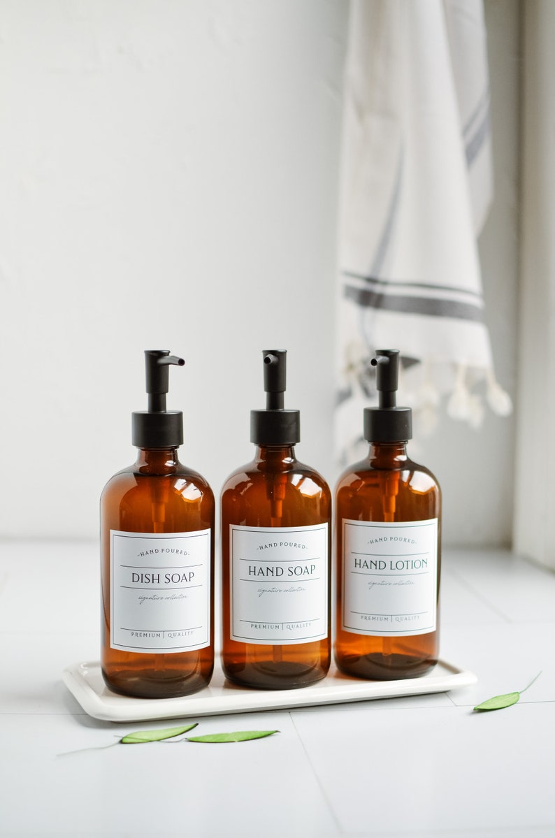 May include: Three amber glass bottles with black pump tops and white labels. The labels read "Hand Poured", "Dish Soap", "Hand Soap", and "Hand Lotion". The bottles are sitting on a white tray.