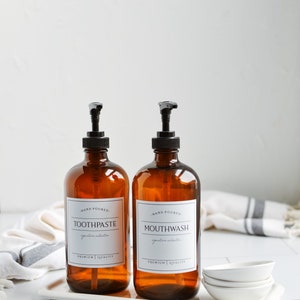 May include: Two amber glass bottles with black pump tops and white labels. The labels read "Hand Poured. Toothpaste. Signature Collection. Premium Quality" and "Hand Poured. Mouthwash. Signature Collection. Premium Quality". The bottles are on a white tray with white ceramic dishes.