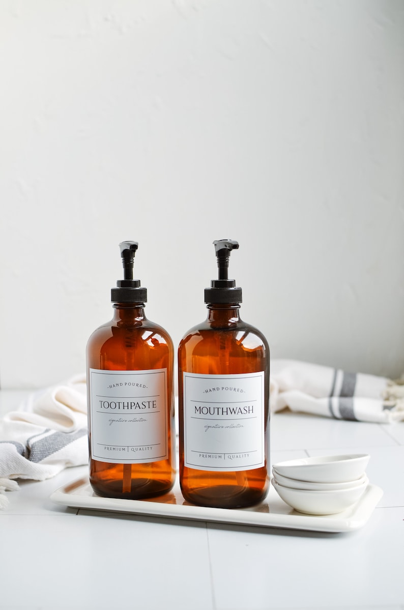 May include: Two amber glass bottles with black pump tops and white labels. The labels read "Hand Poured. Toothpaste. Signature Collection. Premium Quality" and "Hand Poured. Mouthwash. Signature Collection. Premium Quality". The bottles are on a white tray with white ceramic dishes.