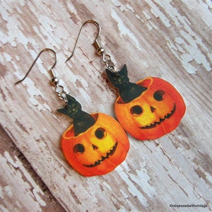 May include: A pair of Halloween-themed earrings featuring black cats peeking out of orange jack-o'-lantern pumpkins.
