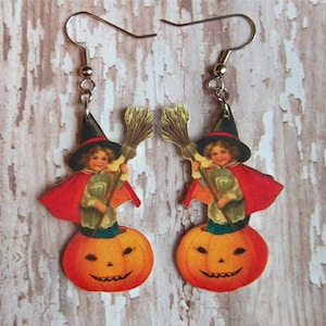 May include: A pair of Halloween-themed earrings featuring a vintage illustration of a child dressed as a witch riding a jack-o'-lantern. The child is holding a broom and wearing a black hat and red cape.