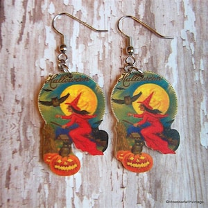 May include: A pair of Halloween-themed earrings featuring a witch flying on a broomstick with a black cat and owls in front of a full moon. The earrings are made of wood and have a vintage look. The text "Halloween" is printed on the earrings.
