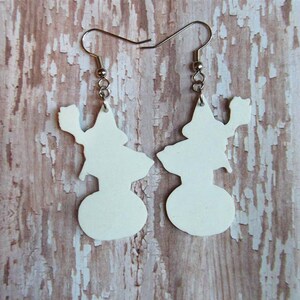May include: A pair of white dangle earrings featuring a silhouette of a witch holding a broom. The earrings are made of a lightweight material and have silver hooks.