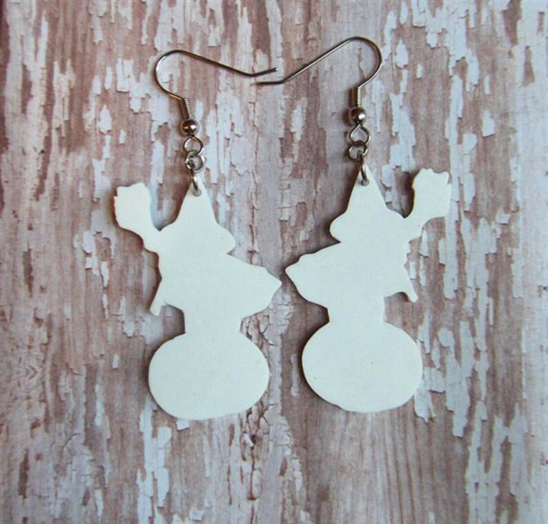 May include: A pair of white dangle earrings featuring a silhouette of a witch holding a broom. The earrings are made of a lightweight material and have silver hooks.