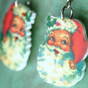 May include: A pair of Christmas-themed earrings featuring a vintage-style image of Santa Claus with a red hat and white beard. The earrings are made of resin and have a glittery finish.