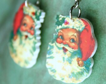 May include: A pair of Christmas-themed earrings featuring a vintage-style image of Santa Claus with a red hat and white beard. The earrings are made of resin and have a glittery finish.