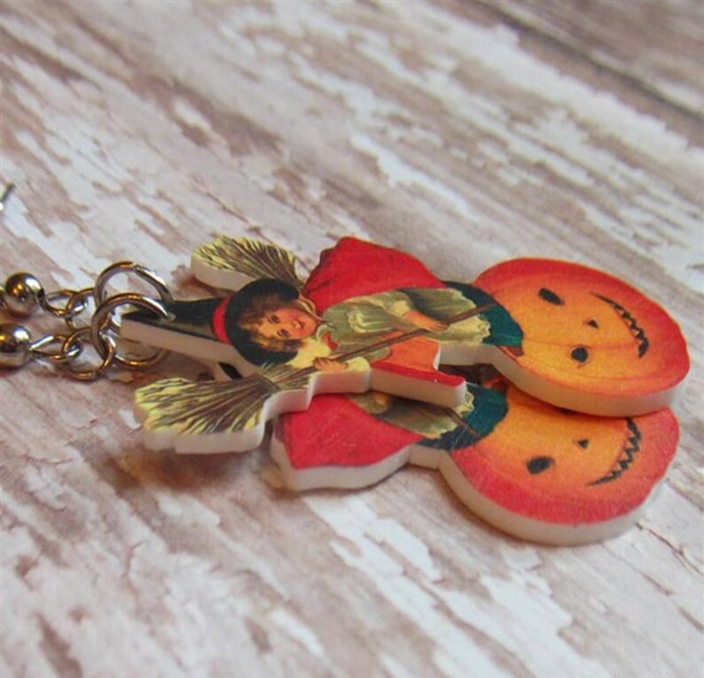 May include: A pair of Halloween-themed earrings featuring a vintage illustration of a witch riding a broomstick with a pumpkin. The earrings are made of white plastic with a red, orange, and yellow color scheme.